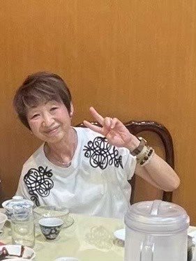 房晴美さん。椅子に座ってピースサインをしている。