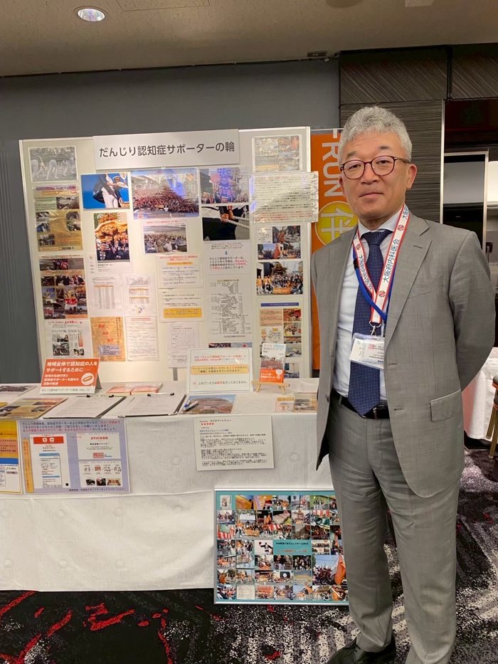 川端徹さん。だんじり認知症サポーターの輪の展示パネルの前に立っている。