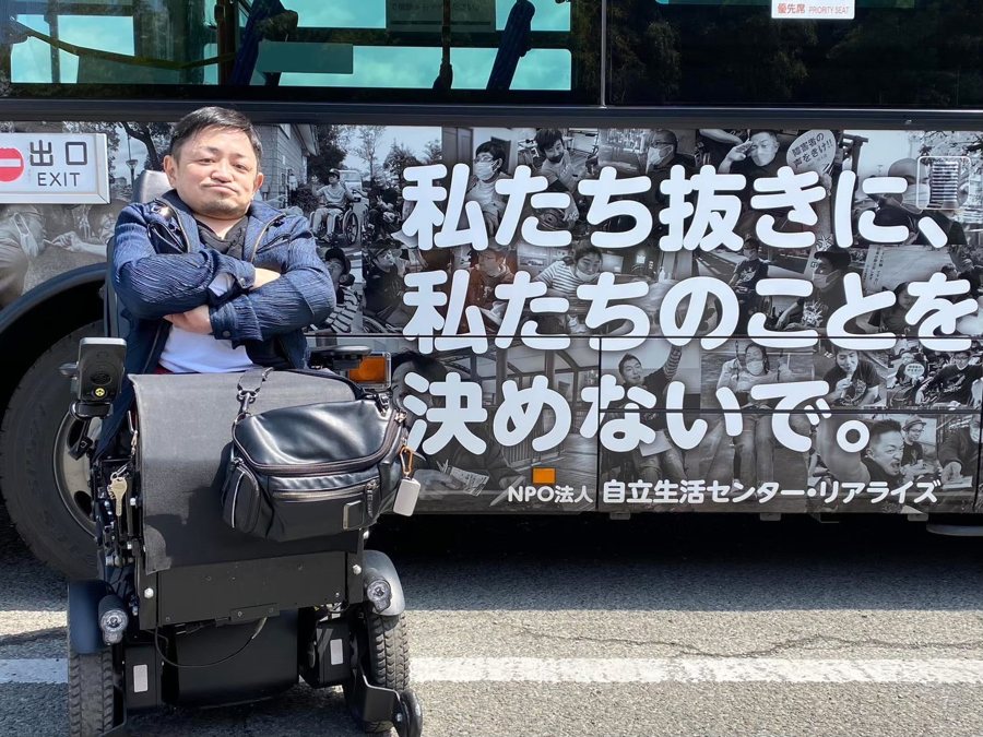 電動車いすの三井孝夫さんが、「私たち抜きに、私たちのことを決めないで。」と書かれたバスの前で腕を組んでいる写真。