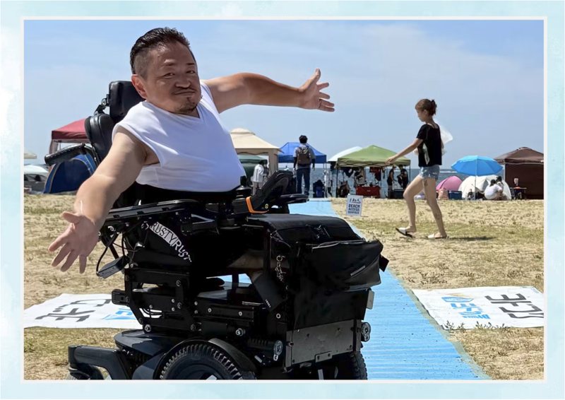 三井孝夫さん。浜辺に敷かれたビーチマットの上で、電動車いすに乗って両手を広げている。
